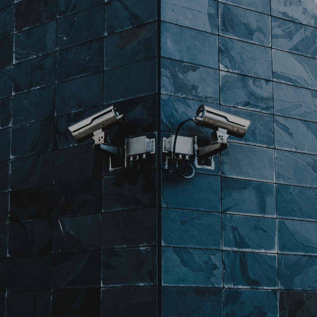 Two surveillance camera on the corner of a dark blue wall monitoring the surrounding areas